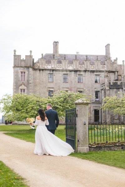 bride and groom walk on the pathway close to grass venue in front she holds bouquet