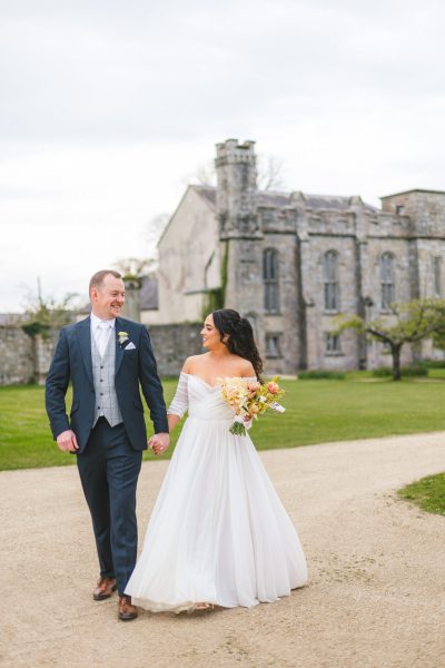 bride and groom walk on the pathway close to grass venue in background she holds bouquet