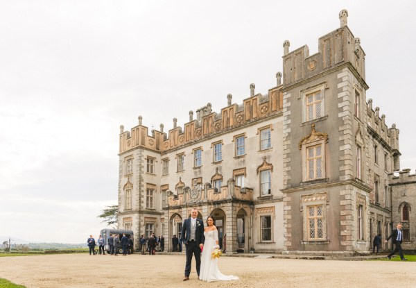 bride and groom walk on the pathway close to grass venue entrance in background she holds bouquet