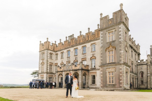 bride and groom walk on the pathway close to grass venue entrance in background she holds bouquet