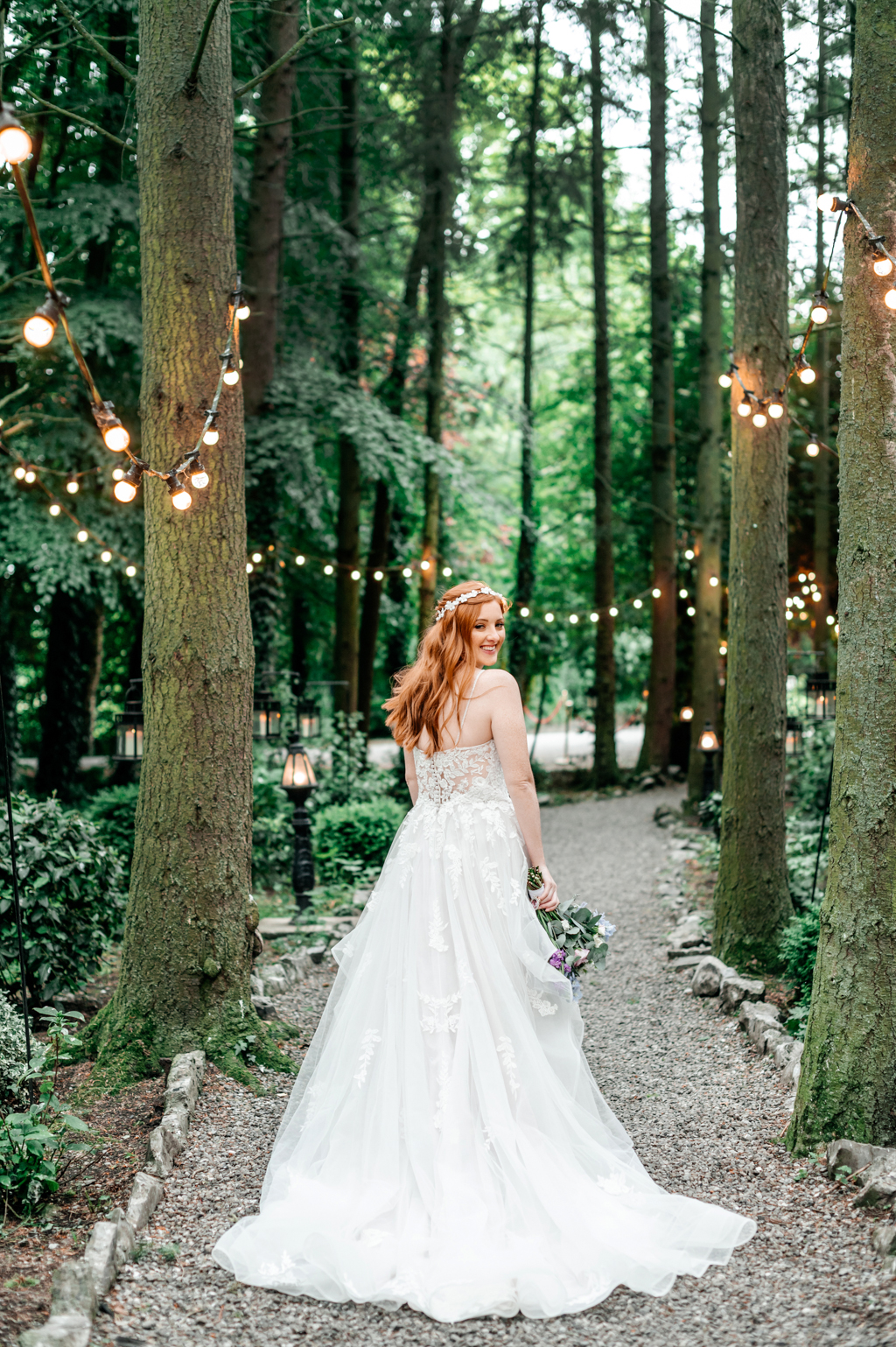 bride stands on pathway to forest setting fairy lights surround