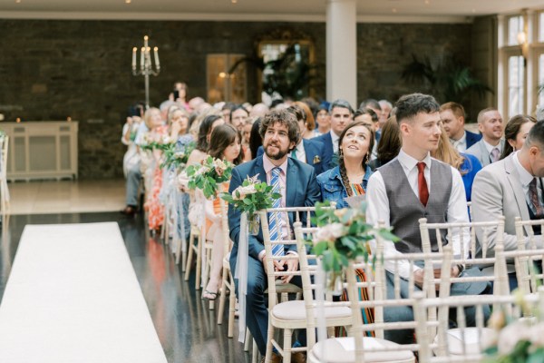 Guests sit in a ceremony hall with pink flowers dotted on end chairs