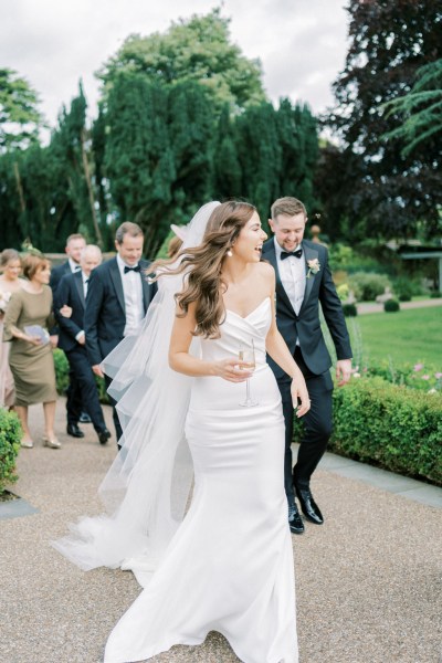 Bride walks along path in garden followed by groom and wedding party