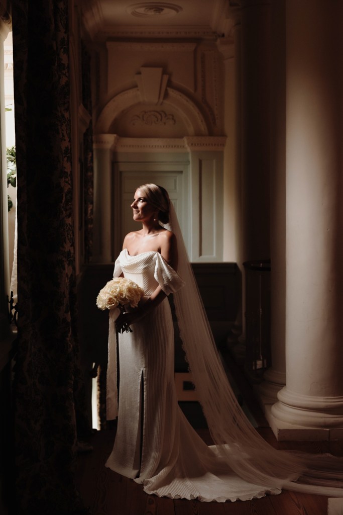 bride stands shadow photographer beside window she holds bouquet