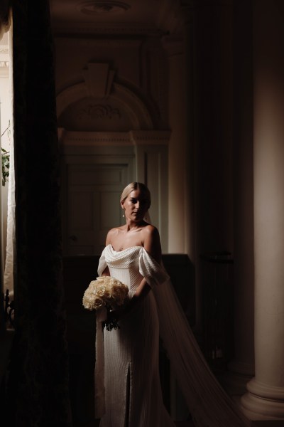 bride stands shadow photographer beside window she holds bouquet