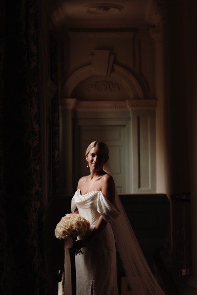 bride stands shadow photographer beside window she holds bouquet