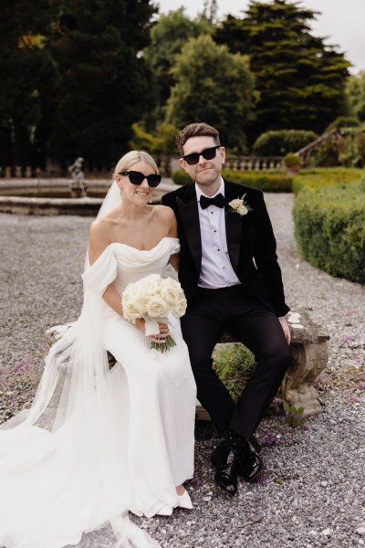 bride and groom are seated in courtyard to wedding venue wearing sunglasses