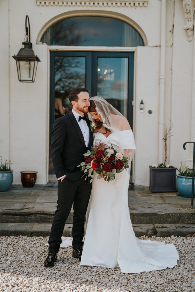 bride and groom pose outside of wedding venue entrance she rests her head on his shoulder