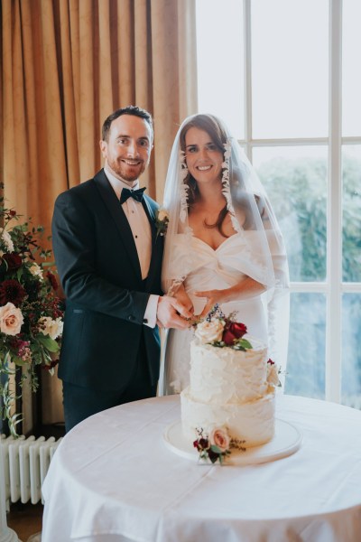 bride and groom cut the white creamy cake that has flowers on top they smile