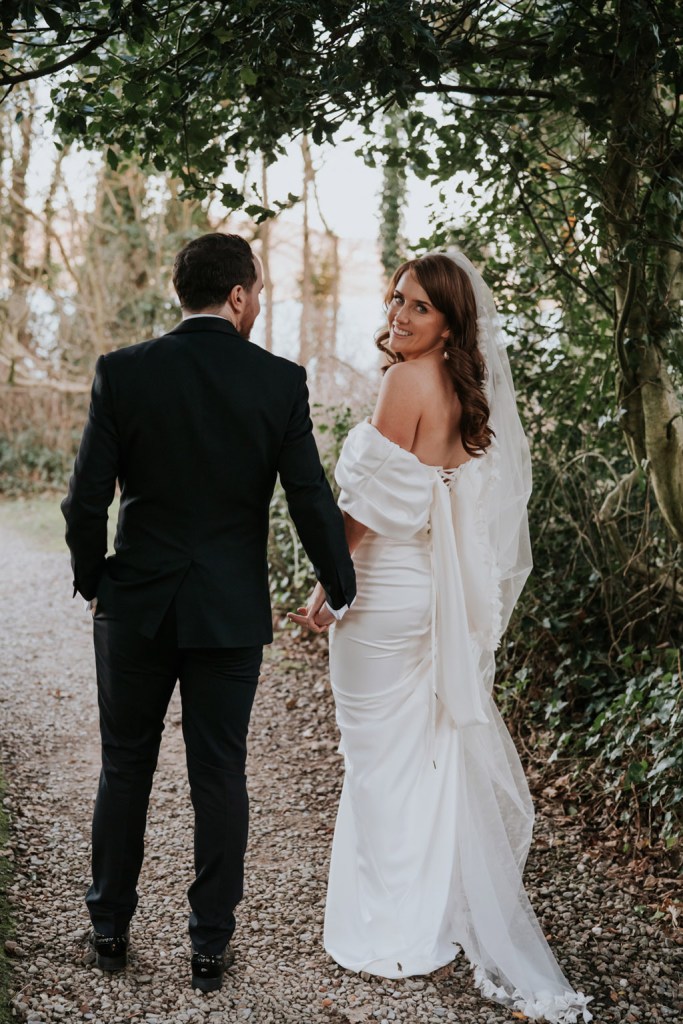 bride and groom walk away from camera on the pathway to forest she looks over her shoulder