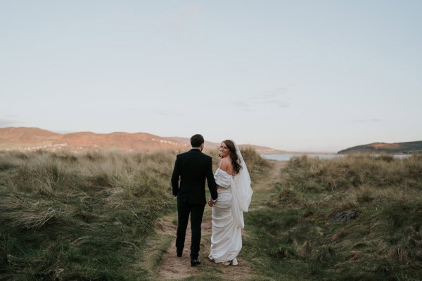 bride and groom walk through the sand and grass landscape they walk away from camera