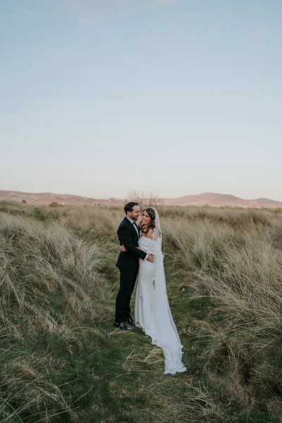 bride and groom walk through the sand and grass landscape they hug