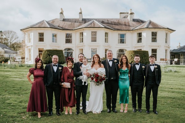 bride groom guests friends mother father pose on the grass in front of wedding venue