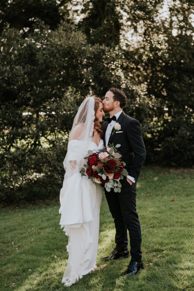 groom kisses bride on foreheads standing on grass to garden