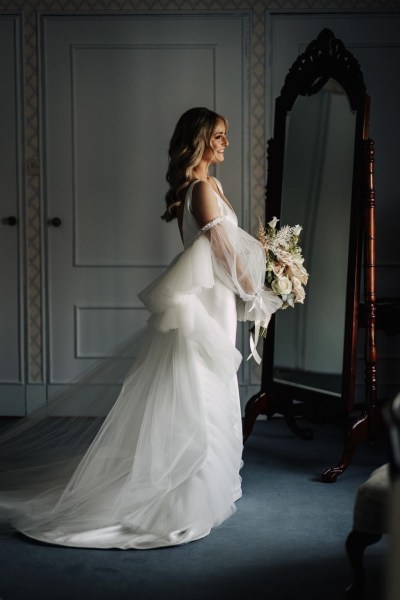 bride stands wedding ready holding bouquet of flowers in front of mirror