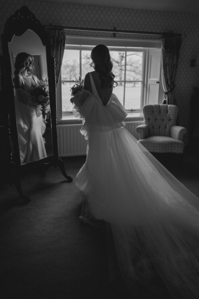 bride stands wedding ready holding bouquet of flowers in front of window in room