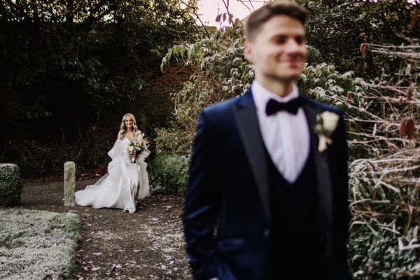 groom waiting for bride to approach him in garden she's holding bouquet