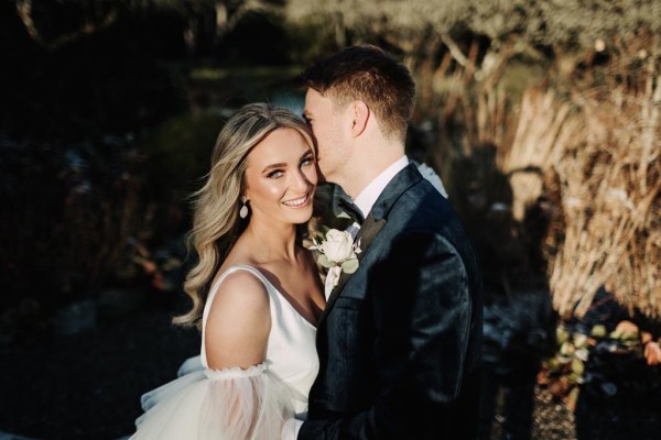 Groom kisses brides cheek in garden park setting as she looks to camera