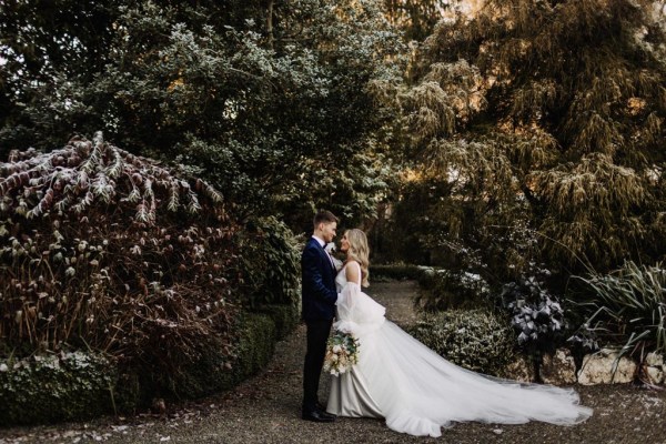 bride and groom are close facing each other she holds bouquet to one side long train in view in garden setting