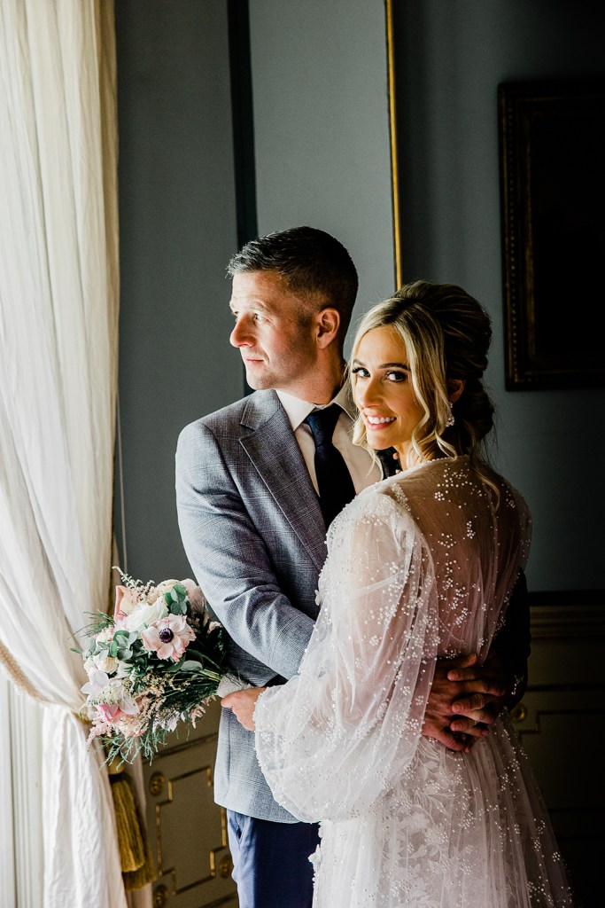 bride and groom stand at window she holds bouquet flowers