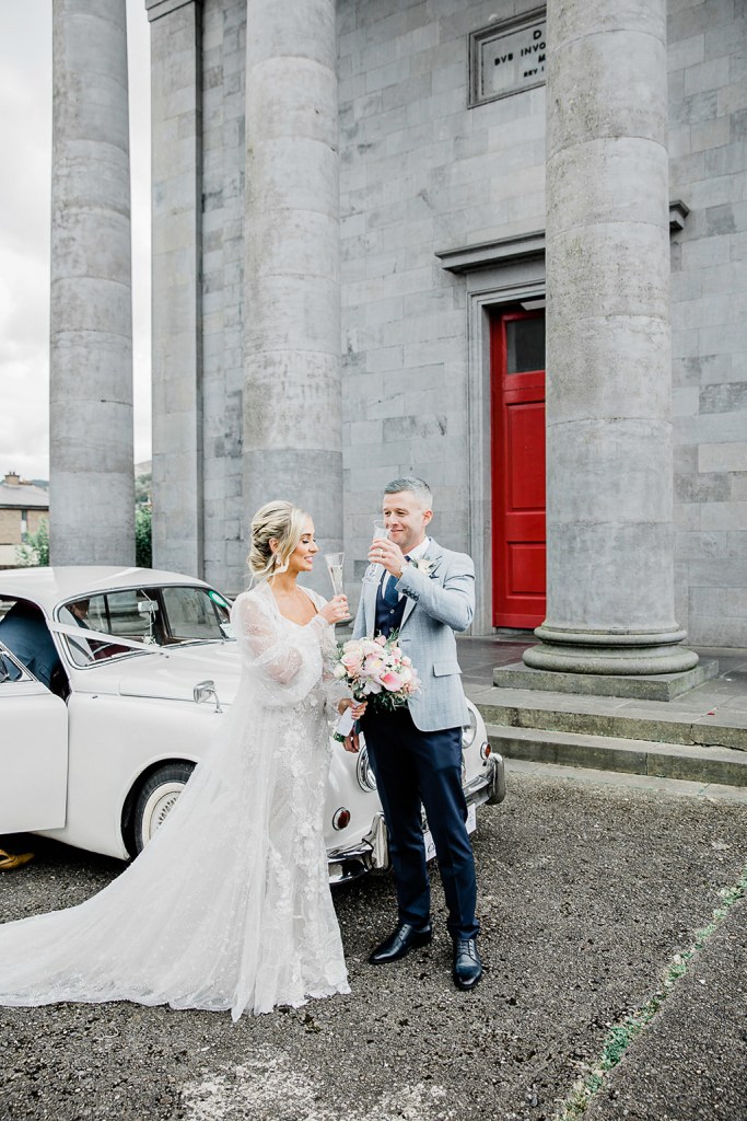 bride and groom stand outside wedding venue against white wedding car she holds bouquet