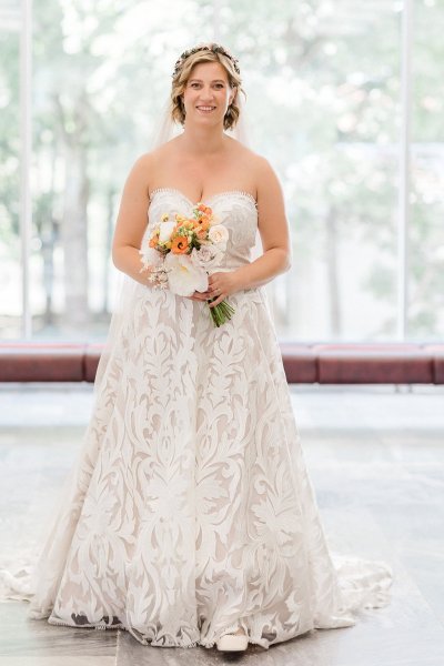 Bride holds orange flowers with bold lace dress before log cabin resort wedding